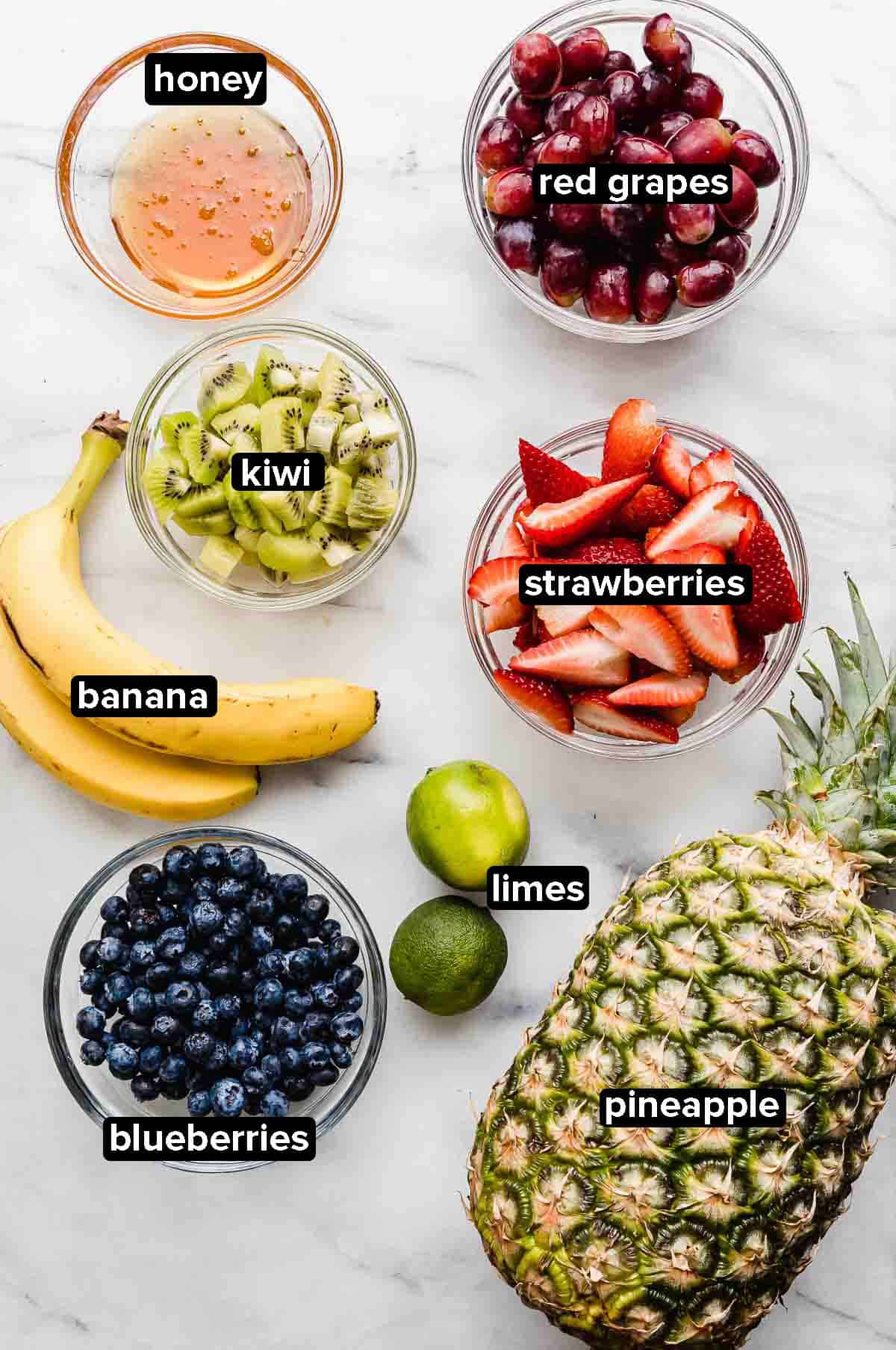 Fruit Cocktail ingredients on a white background: pineapple, kiwi, strawberries, red grapes, limes, honey, bananas, blueberries.