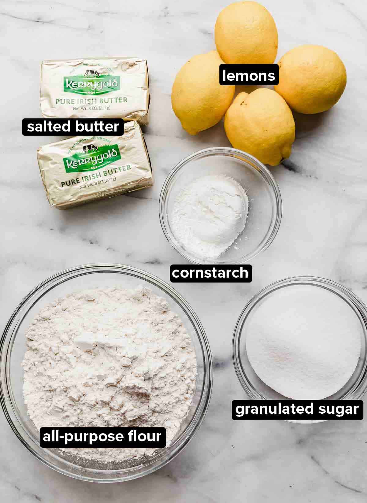 Lemon Shortbread Cookies ingredients portioned into glass bowls on a white and gray marble background: butter, flour, lemons, cornstarch.