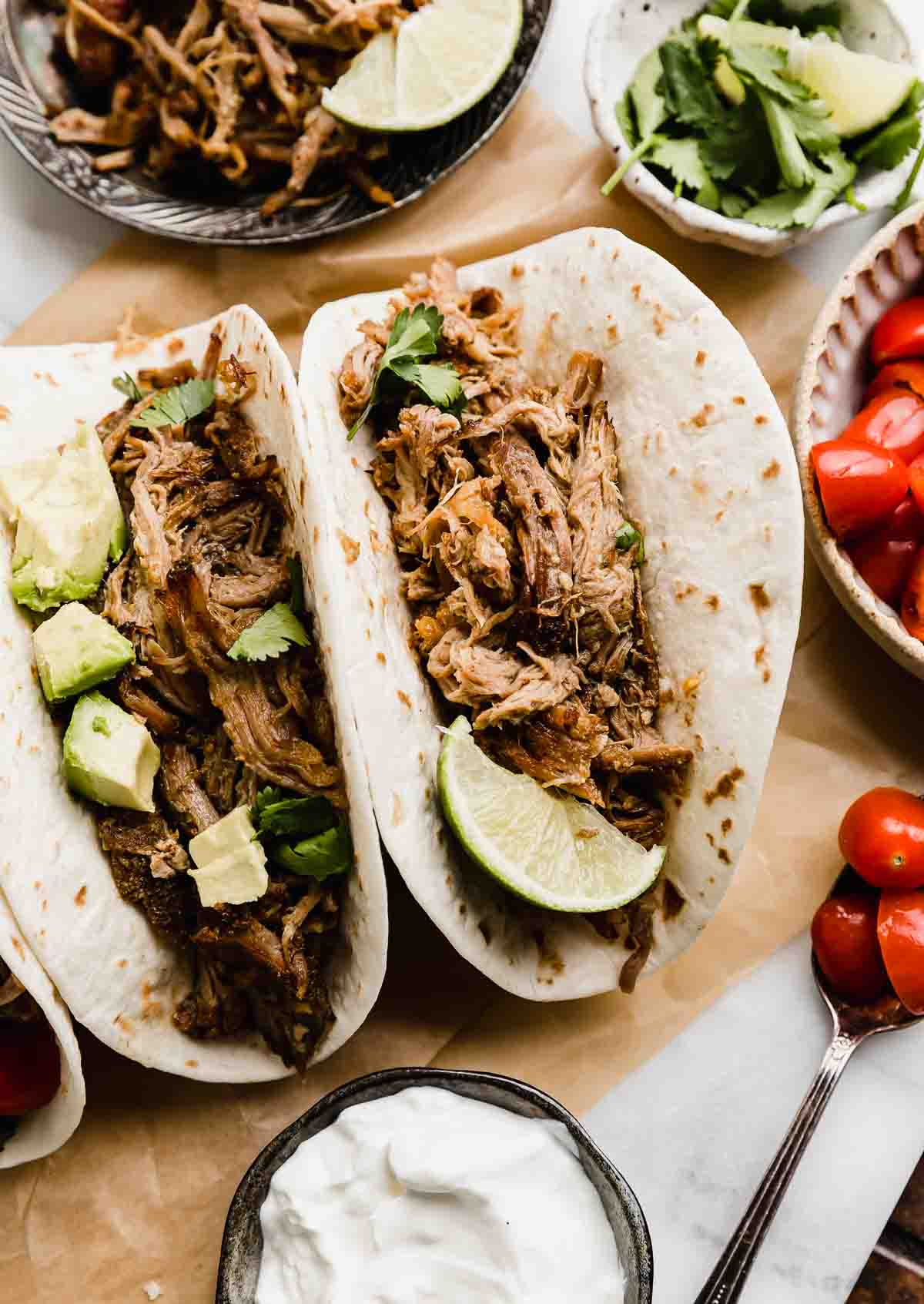 Two flour tortillas filled with slow cooker Mexican pulled pork carnitas, topped with chopped avocado and a side of tomatoes.
