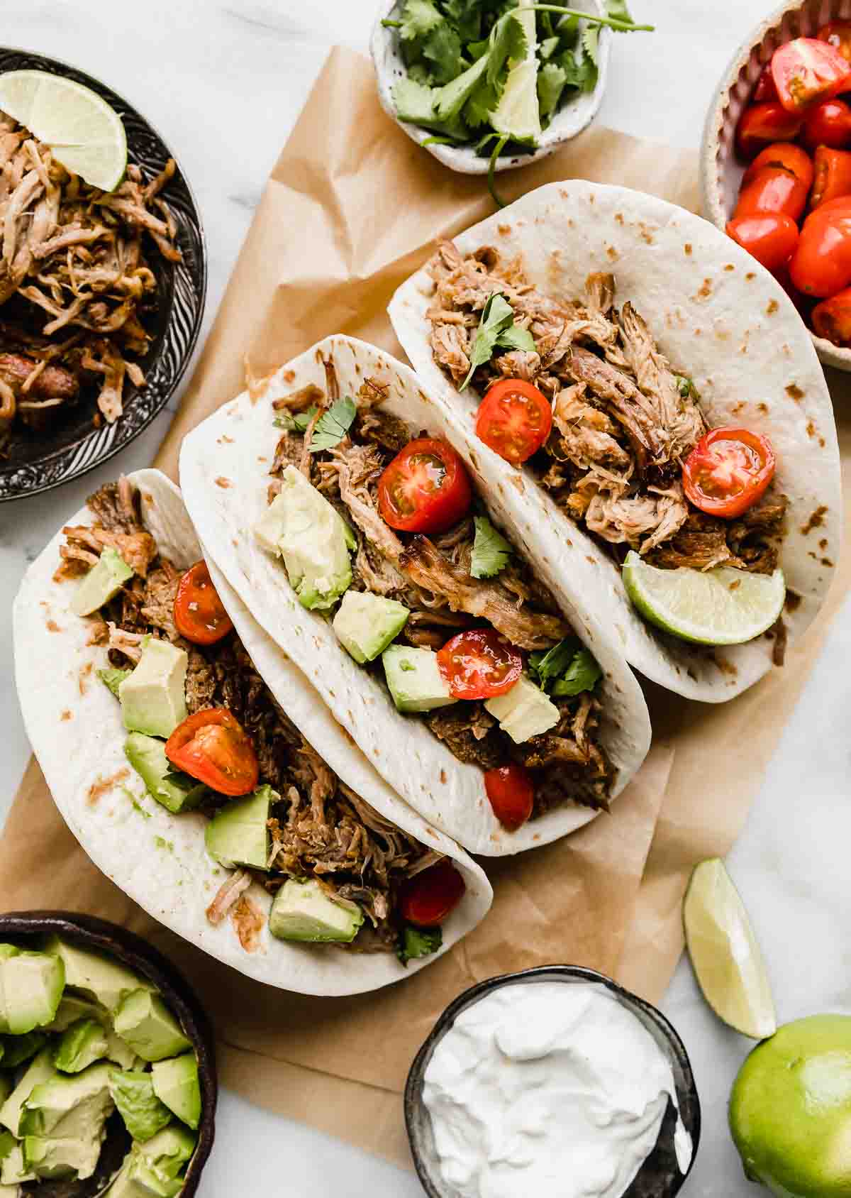 Carnitas on a Kraft colored parchment paper on a white background with avocado and tomatoes surrounding the carnitas.