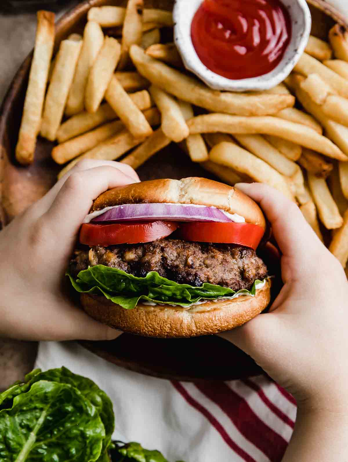 Two hands holding a Steakhouse Burger on a bun with lettuce, tomato, and purple onion, with French fries in the background.
