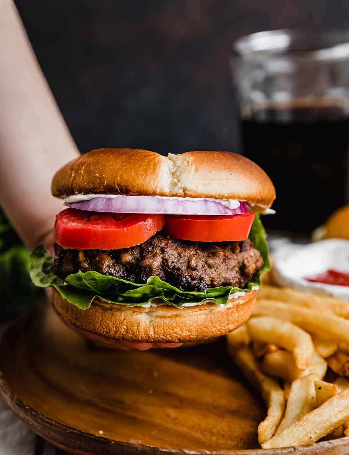 A hand holding up a Steakhouse Burger on a bun with lettuce, tomato, and purple onion, against a dark brown background with French fries in the background.