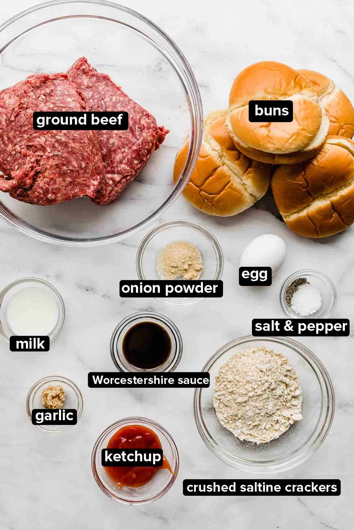 Steakhouse Burgers ingredients on a white marble background: ground beef, buns, ketchup, onion powder, egg, salt and pepper, garlic, milk, and saltine crackers.