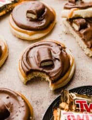 A Twix Cookies made with a shortbread cookie base, topped with caramel, chocolate and a Twix bar on a light brown background.