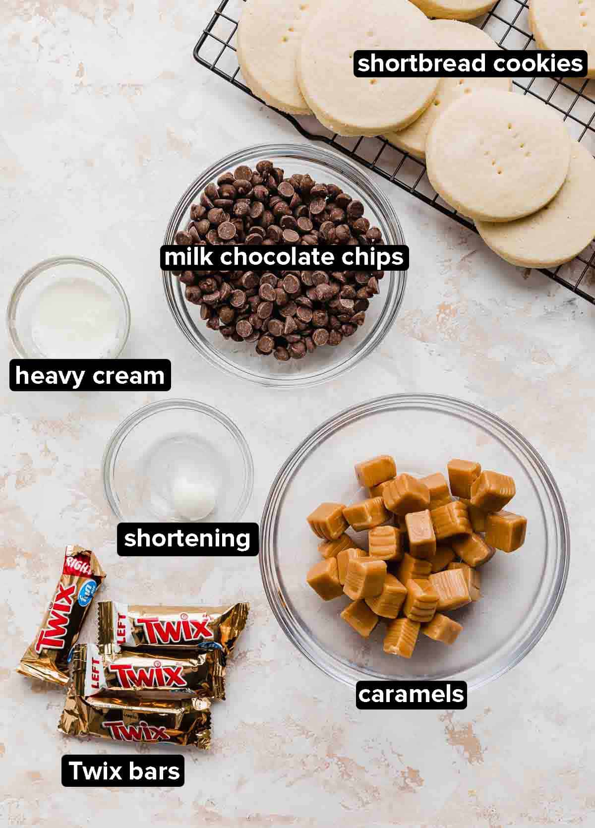 Twix Cookie ingredients on a white and cream textured background: shortbread cookies, caramels, chocolate chips, Twix bars, shortening, and heavy cream.