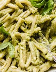 A close up image of pasta tossed in a light green basil pesto with mashed avocado and topped with fresh basil leaves.