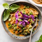 A blue bowl filled with red orange colored Coconut Curry Noodles with vegetables and topped with basil and purple cabbage.