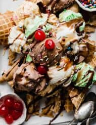 Scoops of chocolate, vanilla, and mint ice cream on top of waffle cones with hot fudge, and maraschino cherries on top.