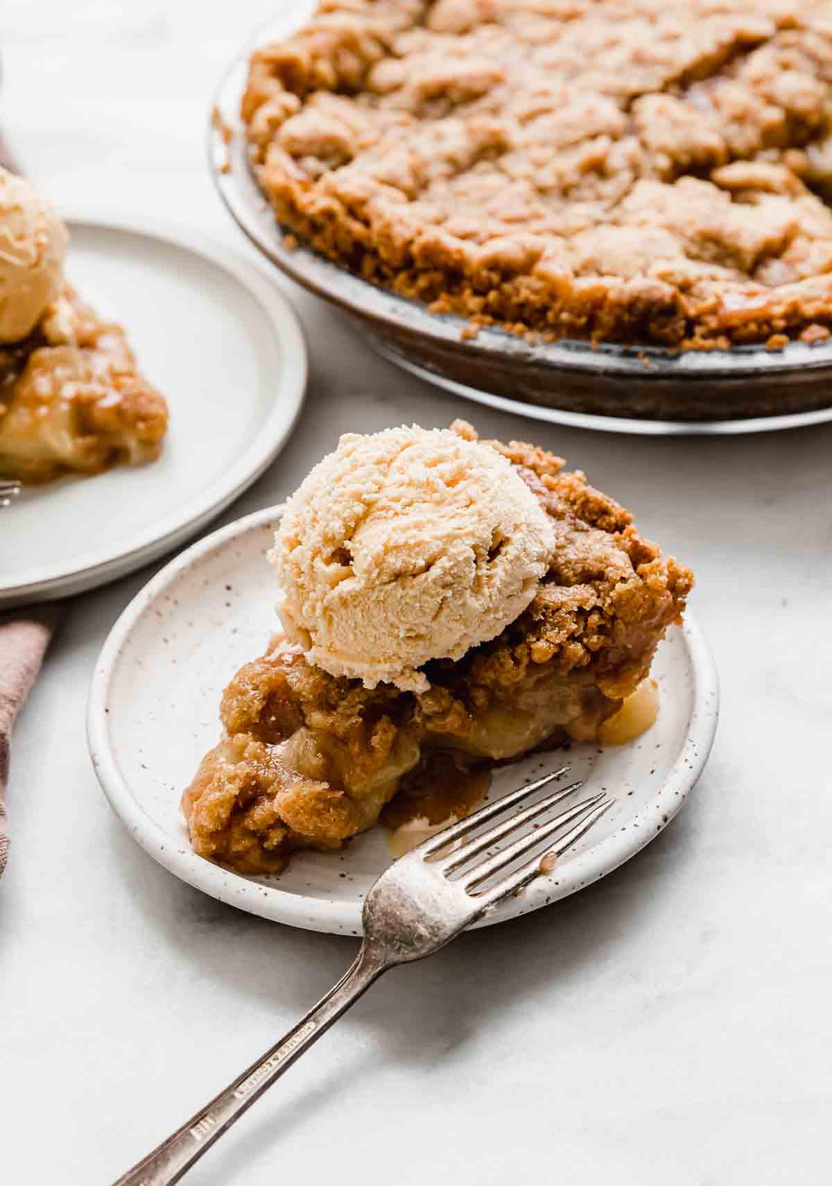 A white plate topped with a slice of Apple Pie baked on a Graham Cracker Crust, topped with a ball of vanilla ice cream.