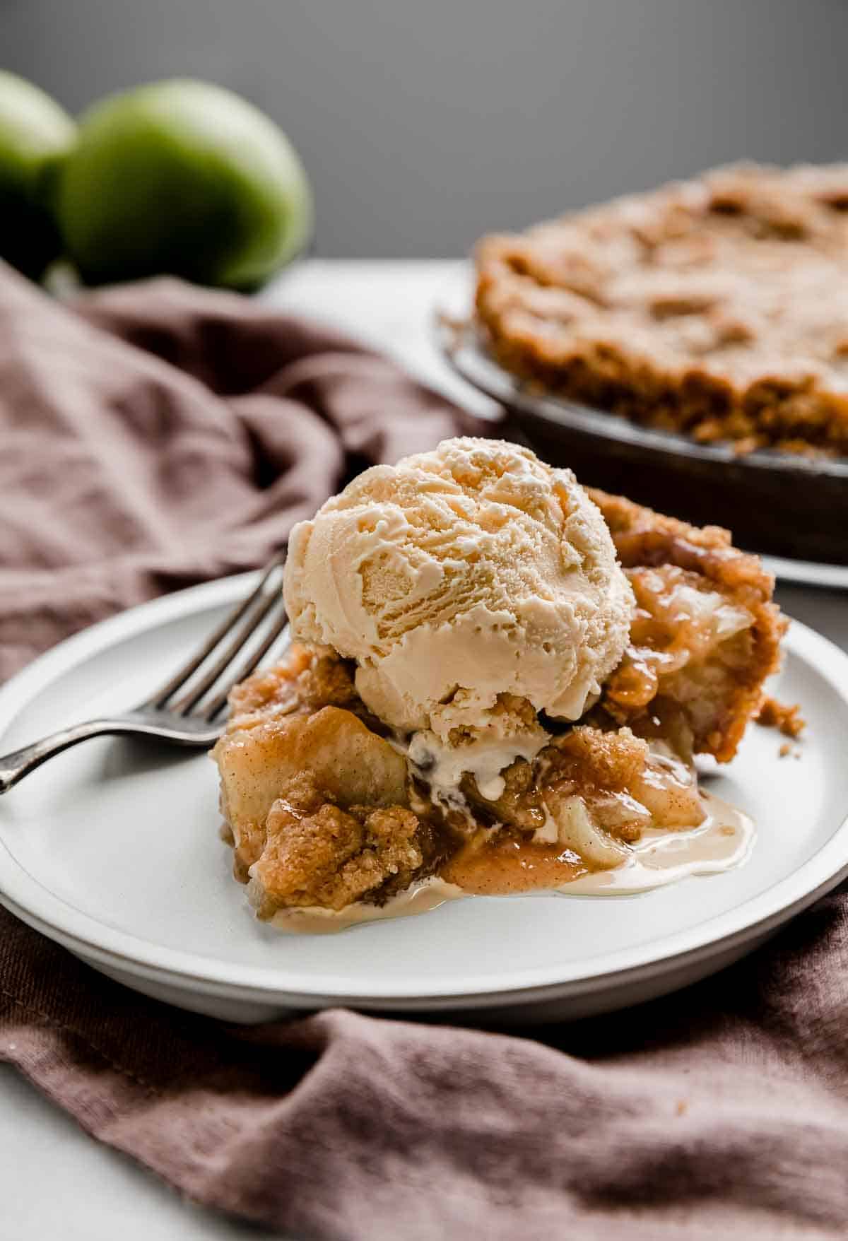 A slice of Apple Pie with Graham Cracker Crust on a white plate with the full apple pie in the background.