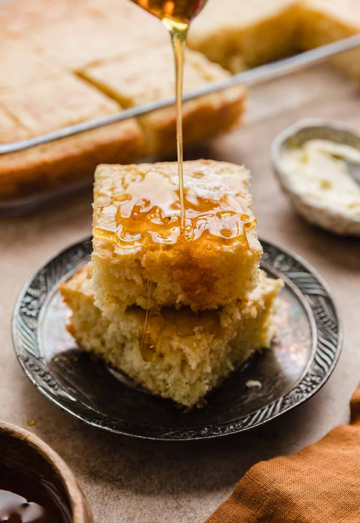 Honey being drizzled over two square slices of Bisquick Cornbread that are sitting on a tarnished plate.