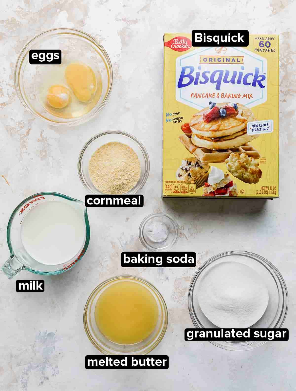 Bisquick Cornbread ingredients on a white and cream textured background; ingredients include a box of yellow Bisquick, cornmeal, milk, eggs, butter, sugar, and baking soda.