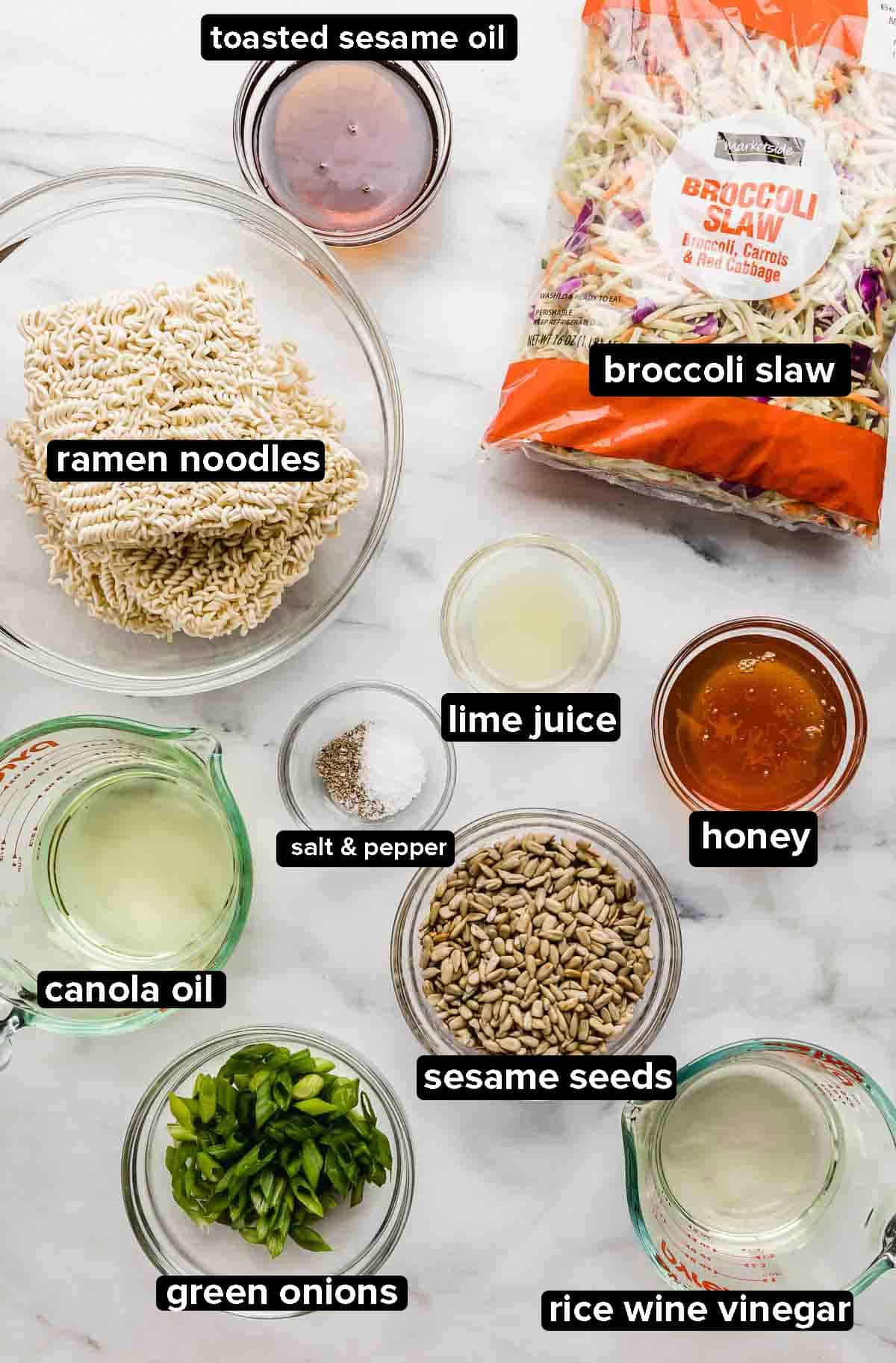 Asian style Broccoli Slaw ingredients on a marble background, including ramen noodles, broccoli slaw, sesame oil, honey, green onions, rice wine vinegar. 