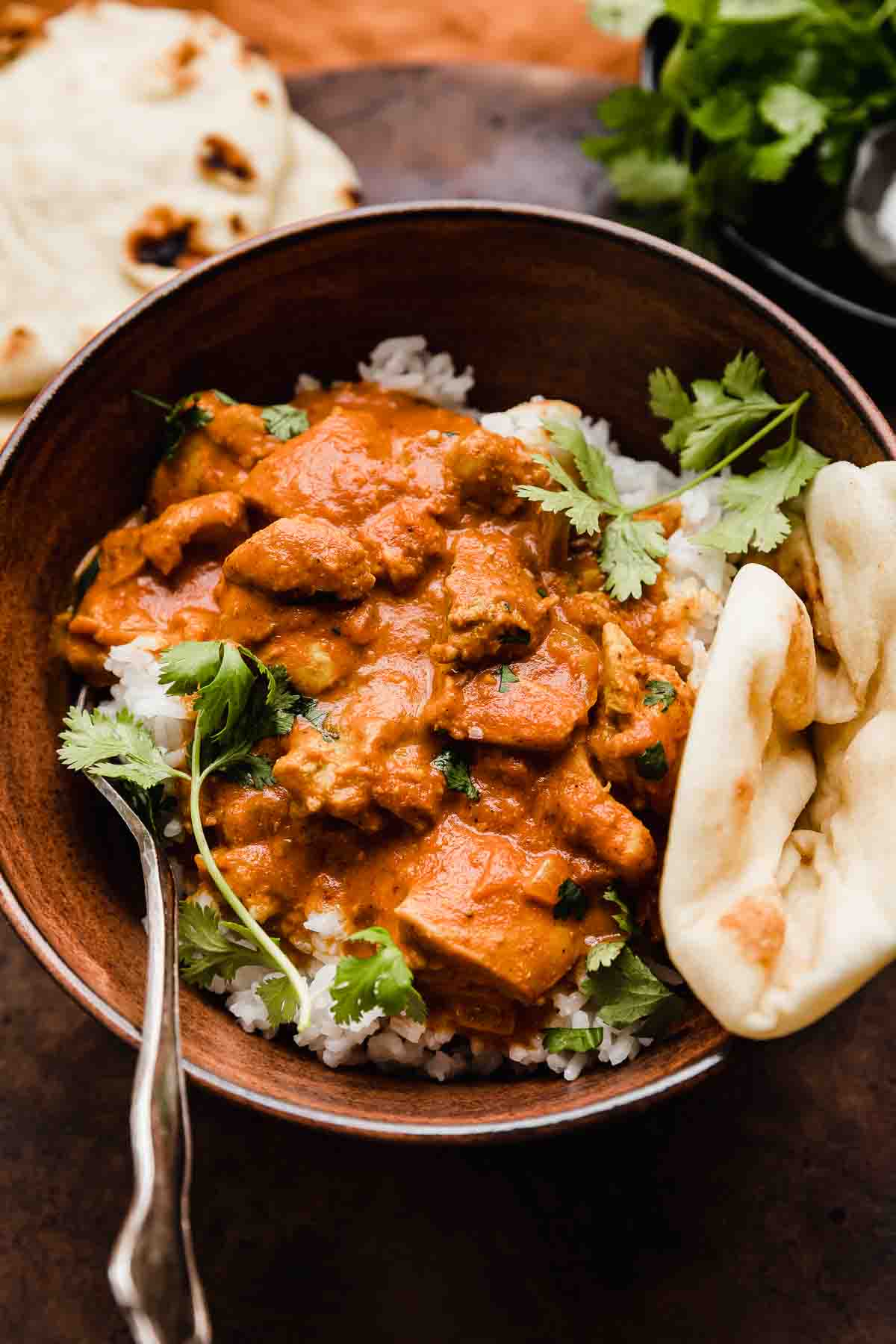 Masala chicken topped with cilantro and a side of naan on a brown background.