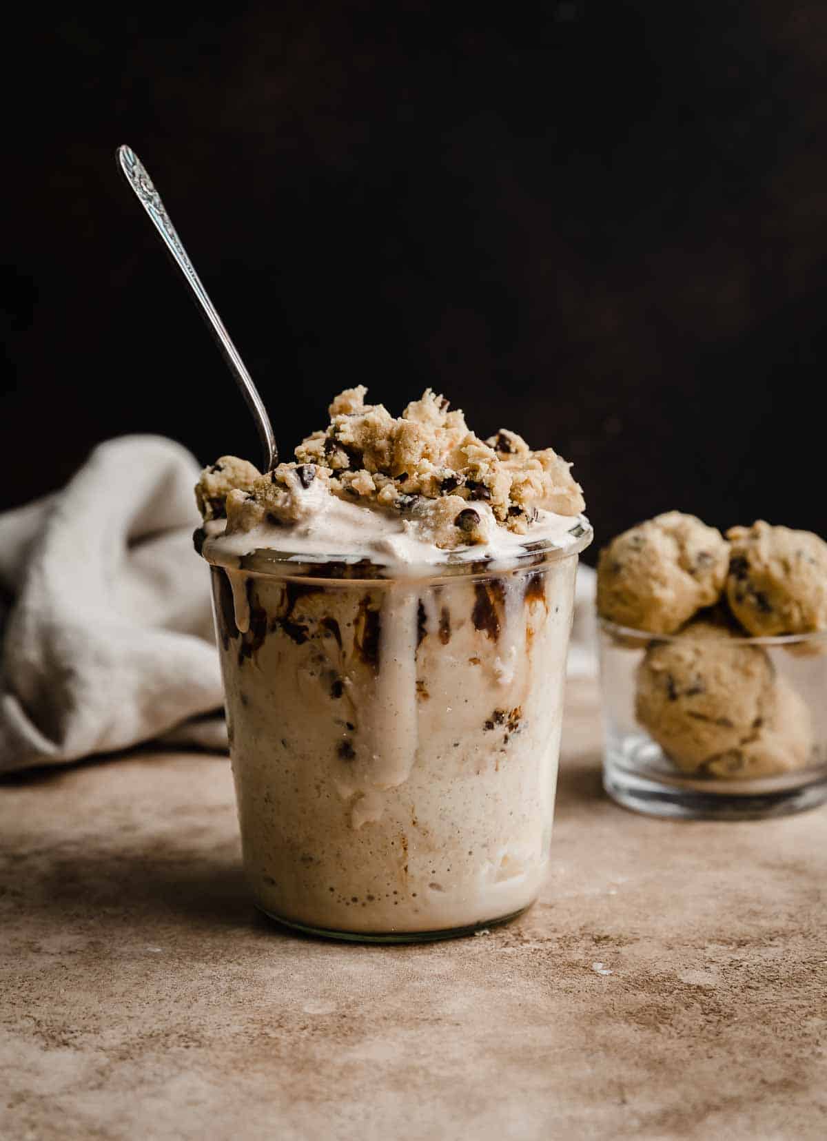A tall glass filled with Chocolate Chip Cookie Dough Blizzard topped with cookie dough chunks, against a brown background.