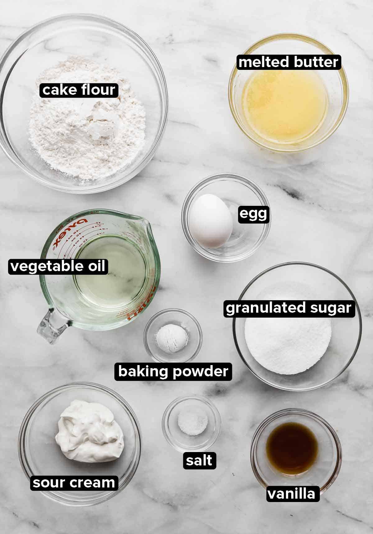 Vanilla Mini Cupcakes ingredients portioned into glass bowls on a white marble background, ingredients include: vegetable oil, cake flour, butter, egg, sugar, sour cream, vanilla, salt, baking powder.