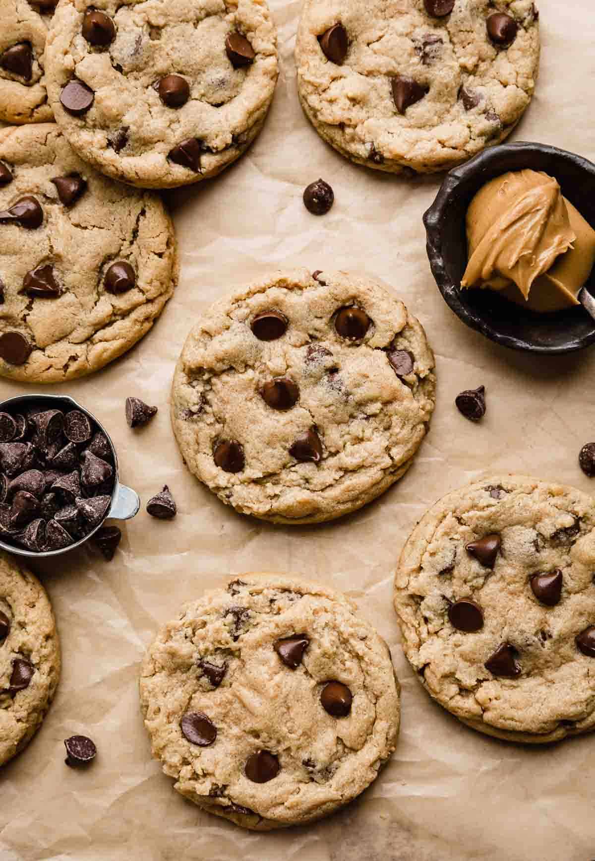 Peanut Butter Chocolate Chip Cookies on a Kraft colored baking parchment paper with chocolate chips surrounding the cookies, and a bowl of peanut butter to the right of the photo.