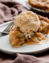 An Apple Pie slice on a white plate topped with a scoop of vanilla ice cream.