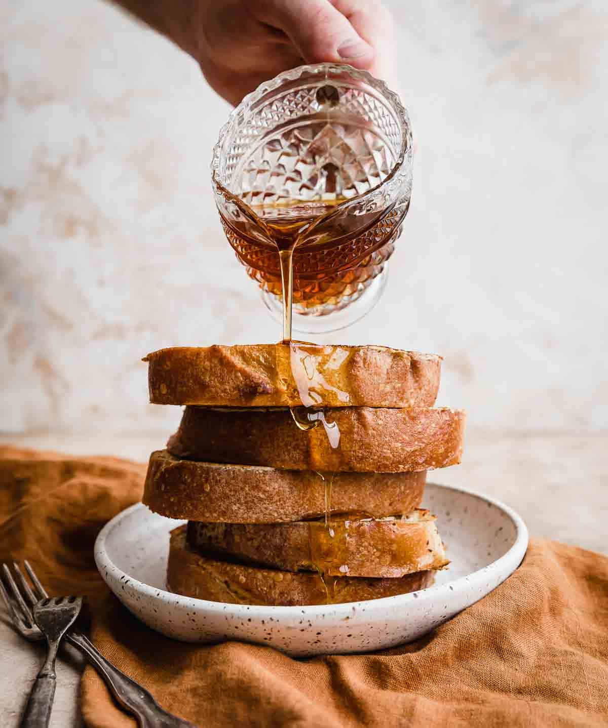 Maple syrup pouring overtop a stack of thick French toast slices.
