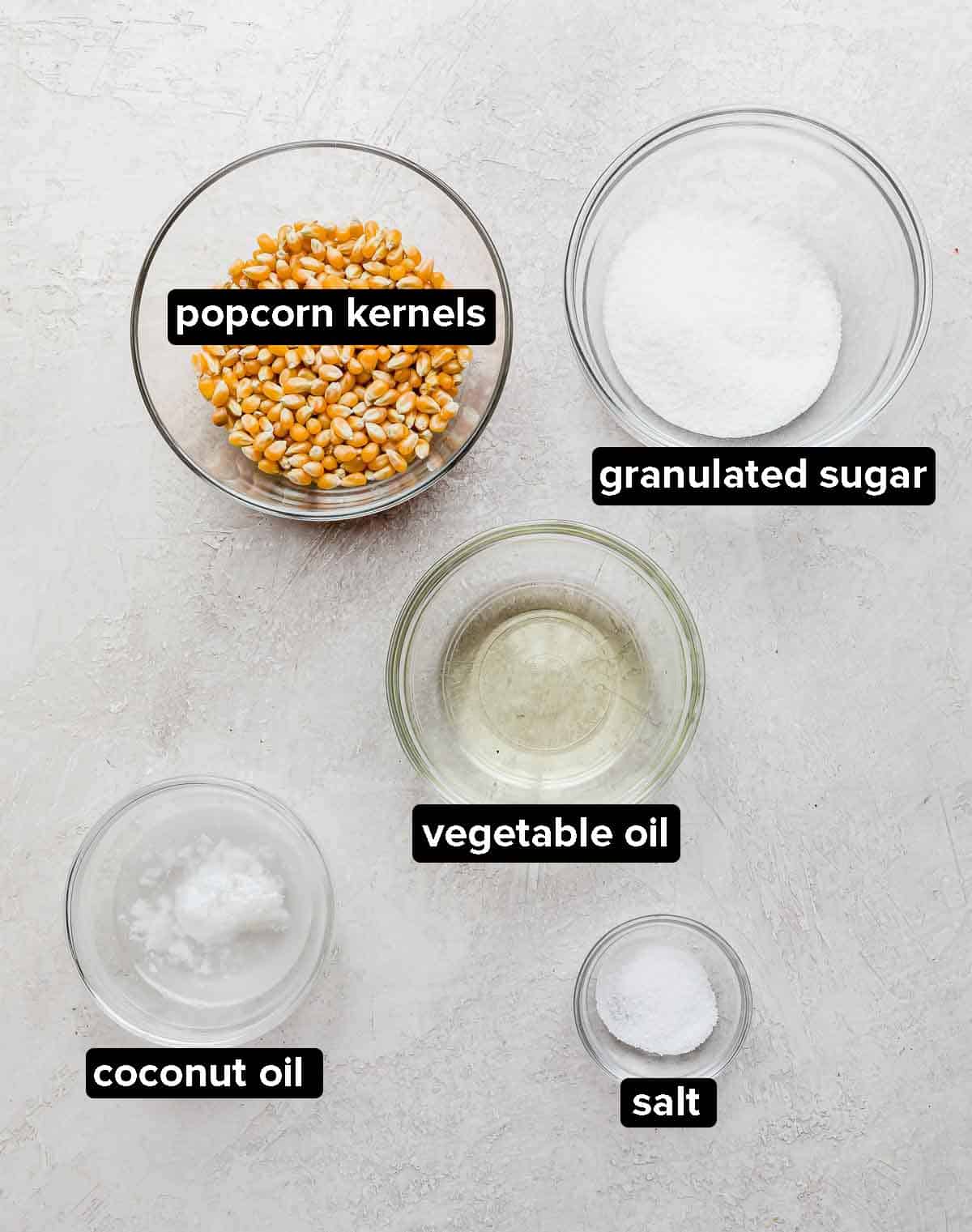Kettle Corn Popcorn ingredients on a light gray textured background: vegetable oil, coconut oil, popcorn kernels, salt, and granulated sugar.