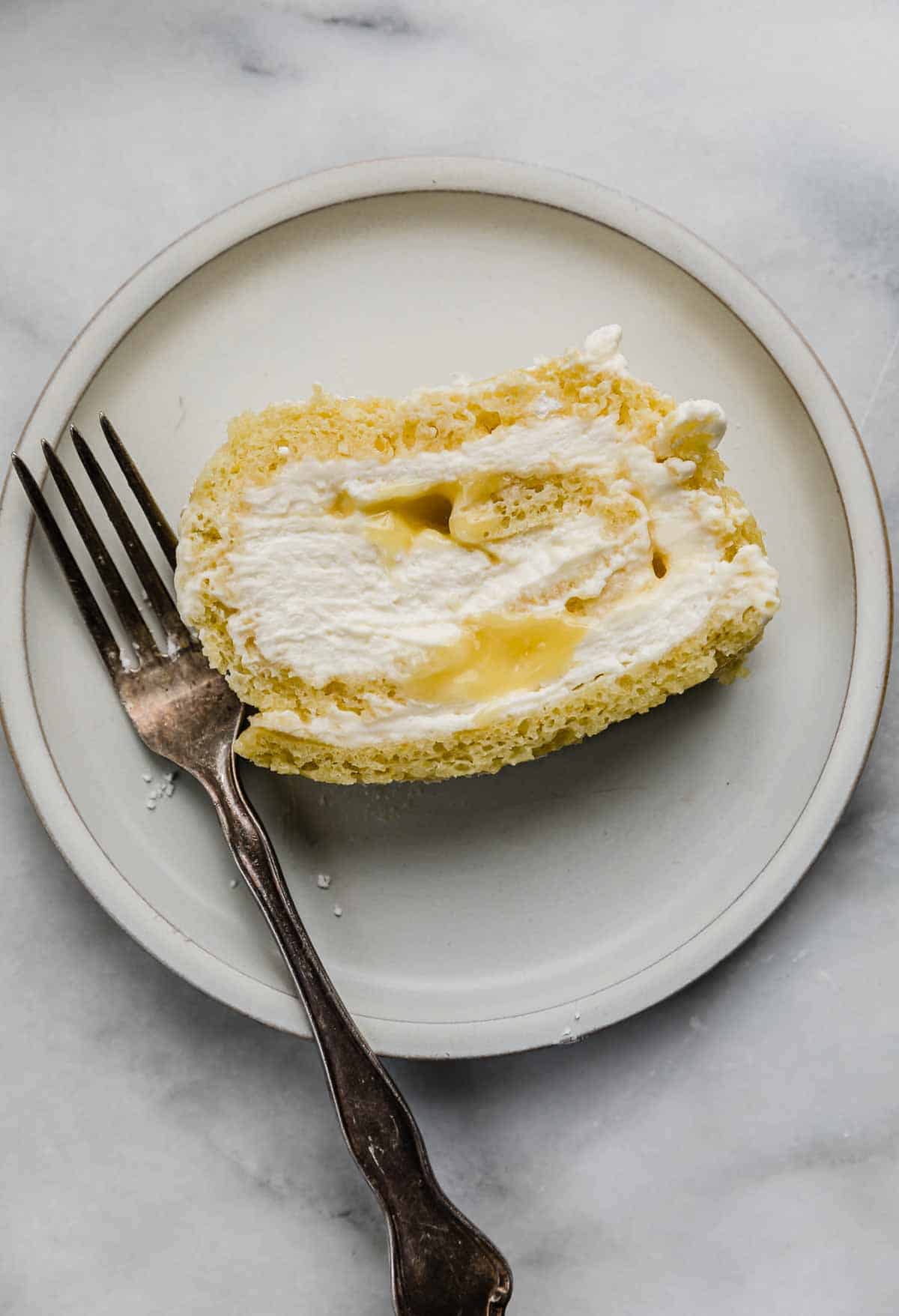 A slice of Lemon Swiss Roll filled with cream and lemon curd, on a white plate with a fork to the left of the cake.