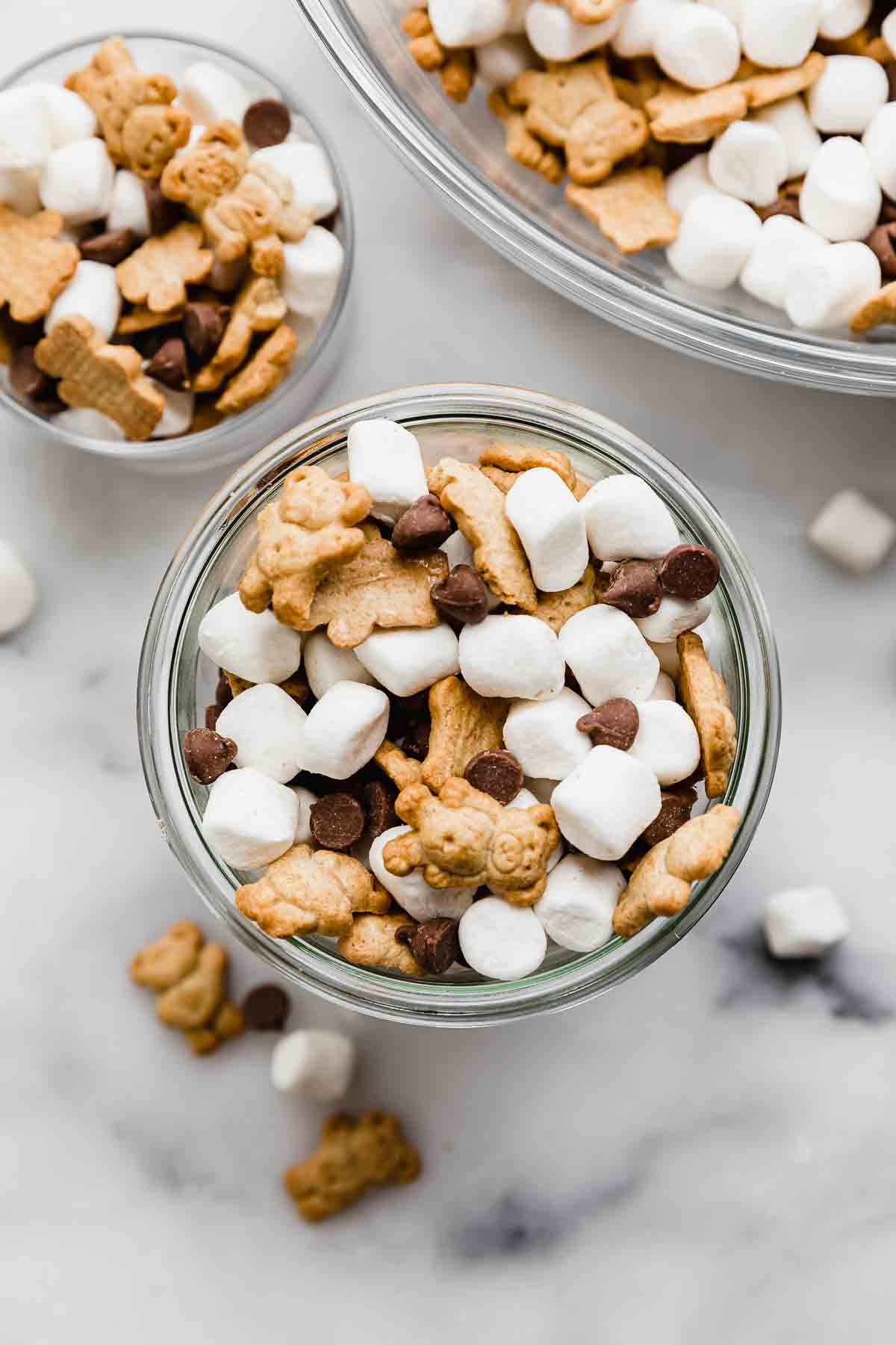 S'mores Mix in a glass cup on a white marble background, the S'mores trail mix consists of mini marshmallows, teddy grahams, and milk chocolate chips.