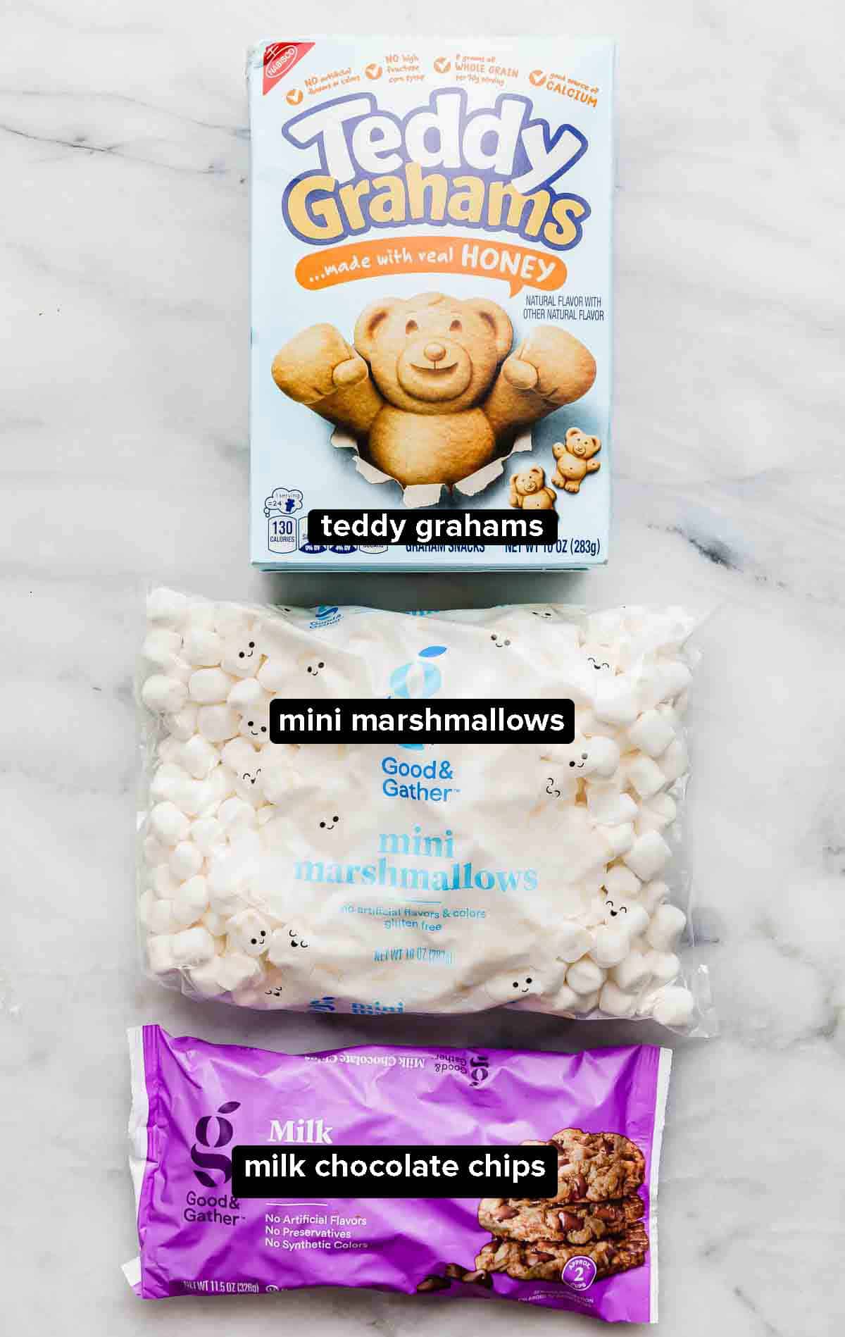 S'mores snack Mix ingredients: a box of teddy grahams, mini marshmallows, and a bag of milk chocolate chips on a white background.