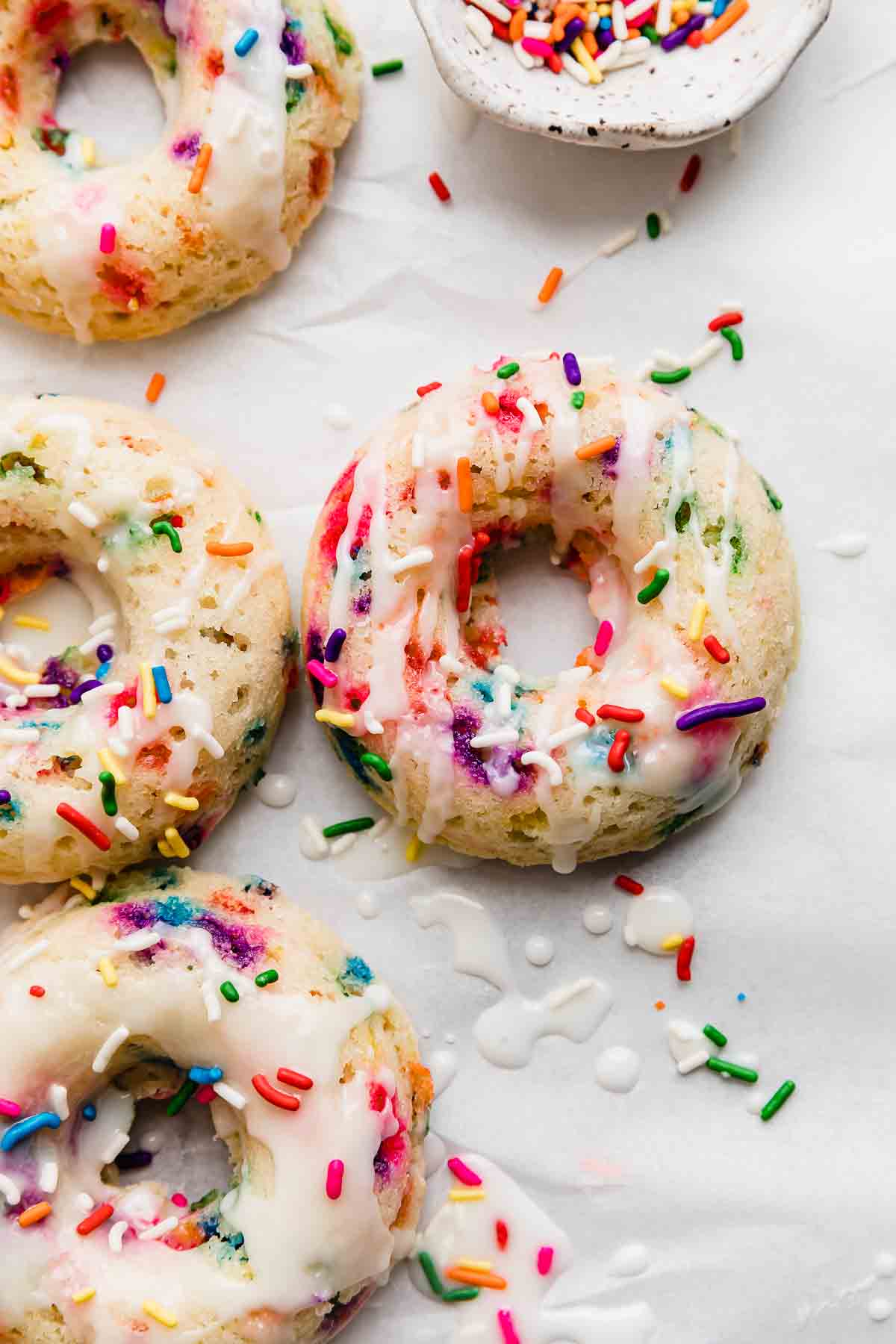 Four Sprinkle Donuts topped with a glaze and colorful sprinkles on a white background. 