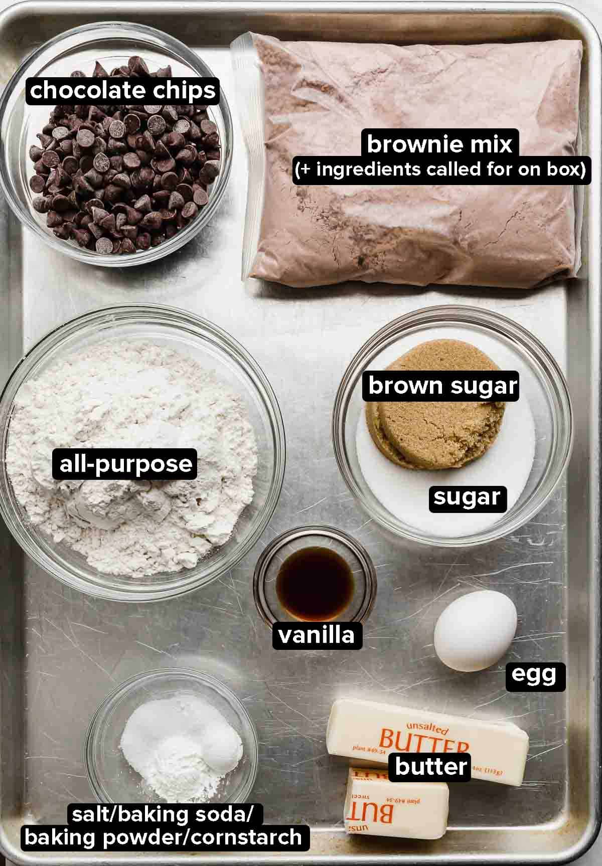 Brookies ingredients on a silver baking sheet: brownie mix, chocolate chips, flour, sugar, butter, leavening and egg.