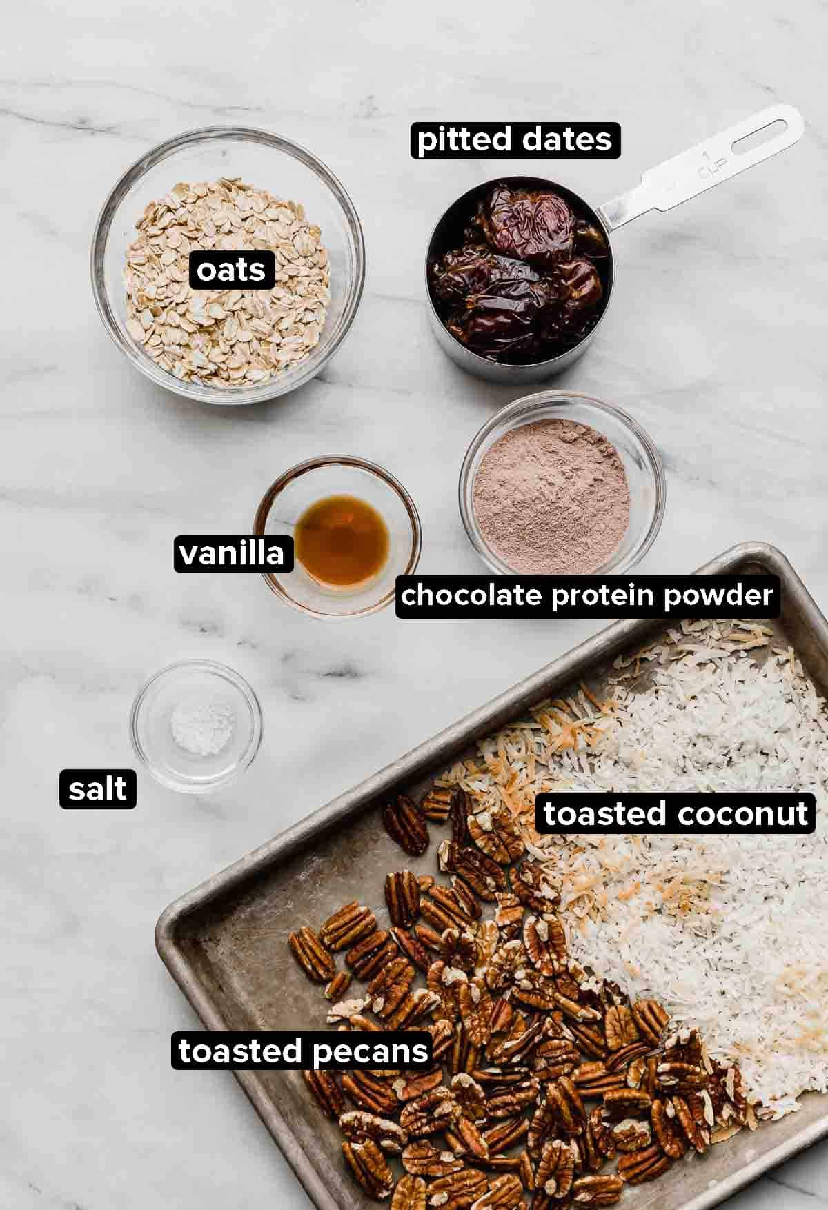 German Chocolate protein balls ingredients on a white marble background: pecans, coconut, dates, oats, vanilla, and protein powder.