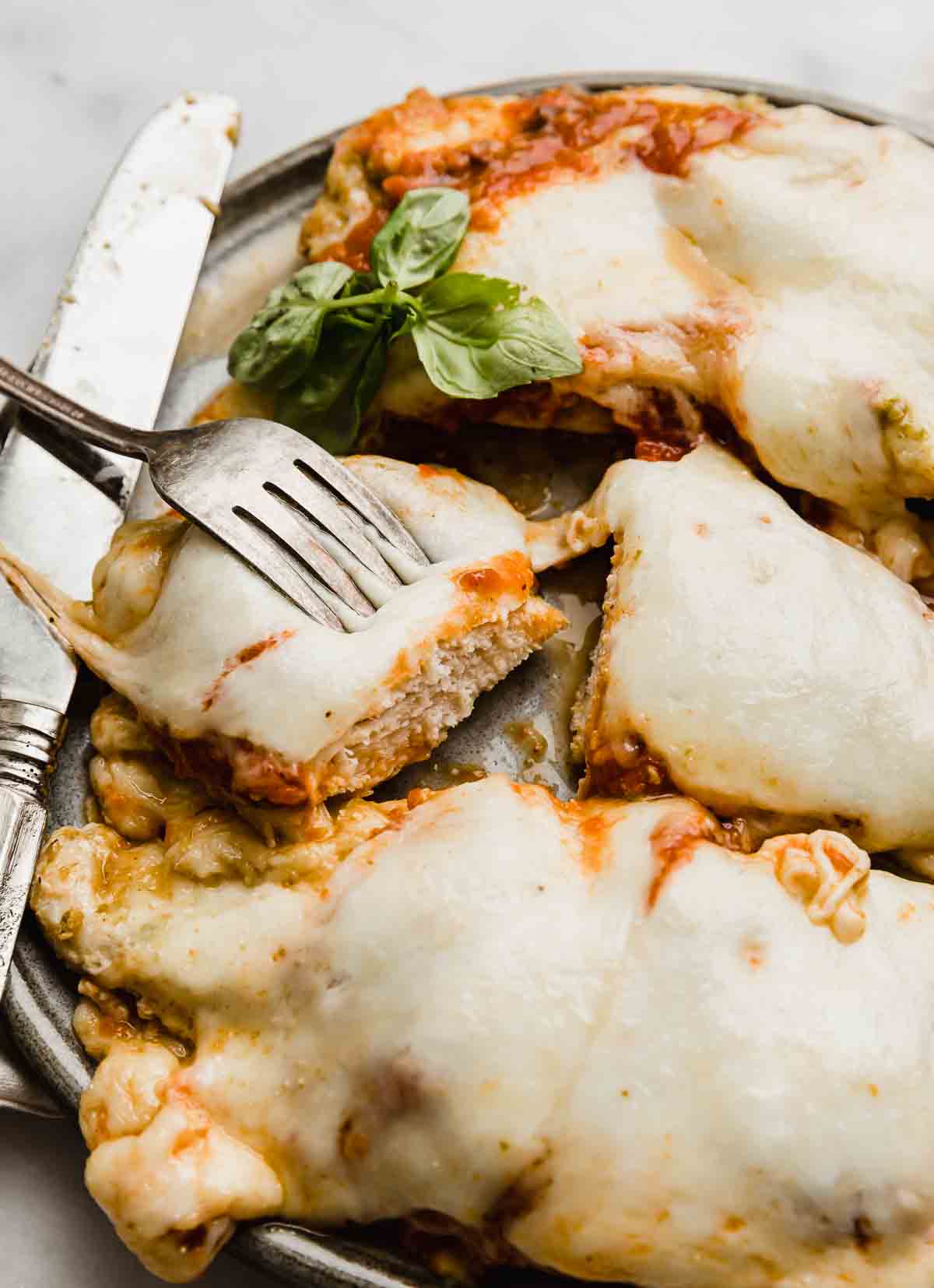 A fork holding up a piece of Pesto Chicken Parmesan over a gray plate.