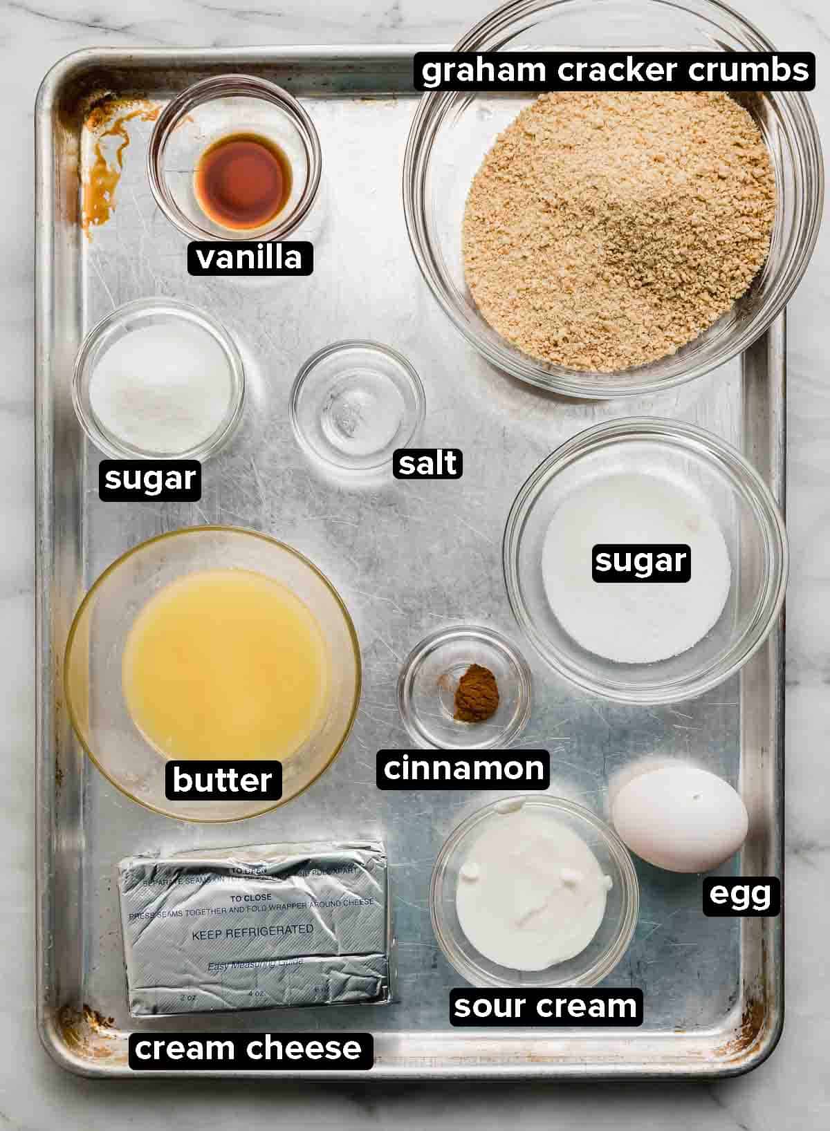 Ingredients used to make the best Mini Cheesecakes recipe portioned into glass bowls on a silver baking sheet: graham cracker crumbs, butter, sugar, egg, vanilla, and cream cheese.