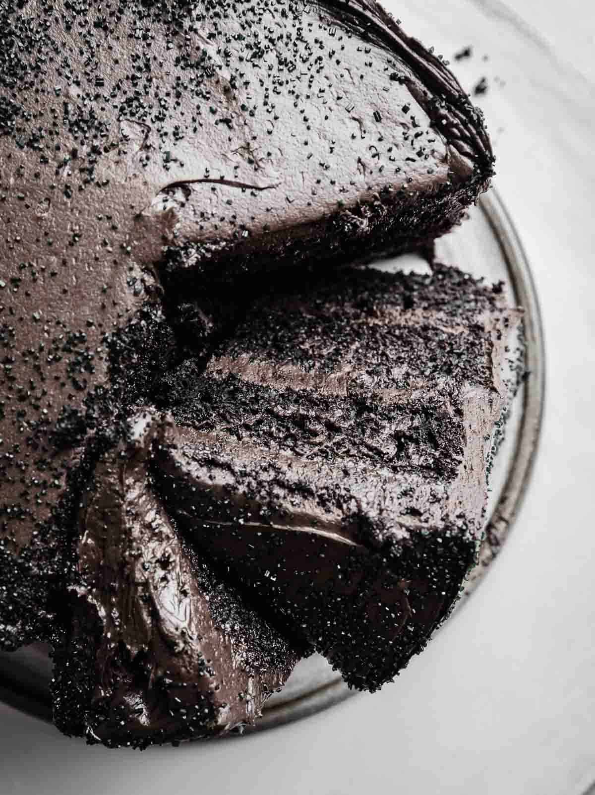 Three layered black velvet cake topped with black frosting, with two slices leaning against each other on a white background.