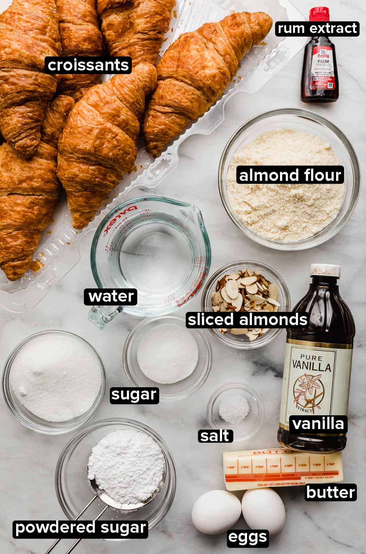 Almond Croissants recipe ingredients on a white background: croissants, almond flour, water, vanilla, eggs, butter, sugar, sliced almonds, and rum extract.