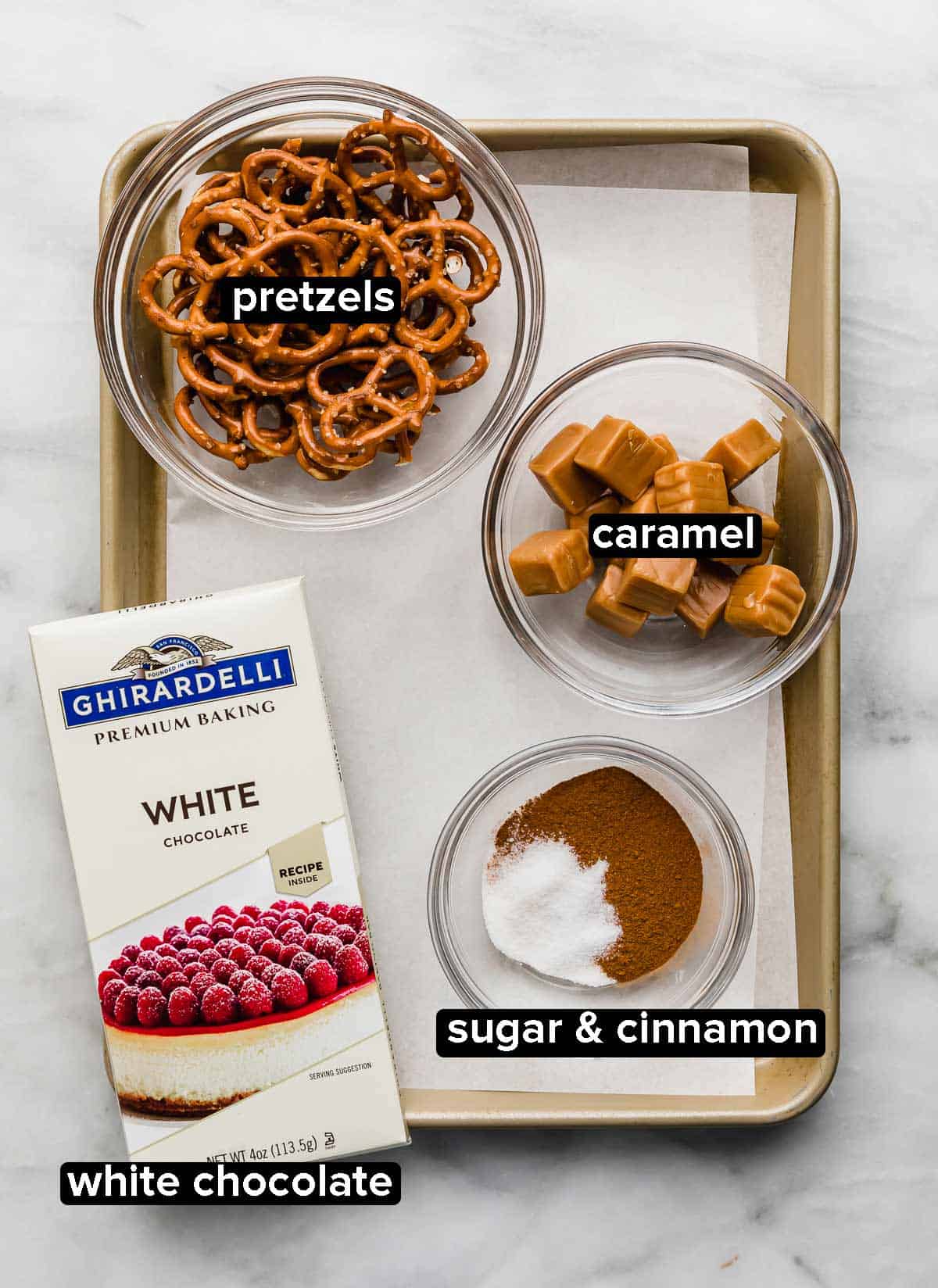 A baking sheet with white chocolate bar, pretzels, caramel, and cinnamon sugar on it, used to make snickerdoodle pretzels.