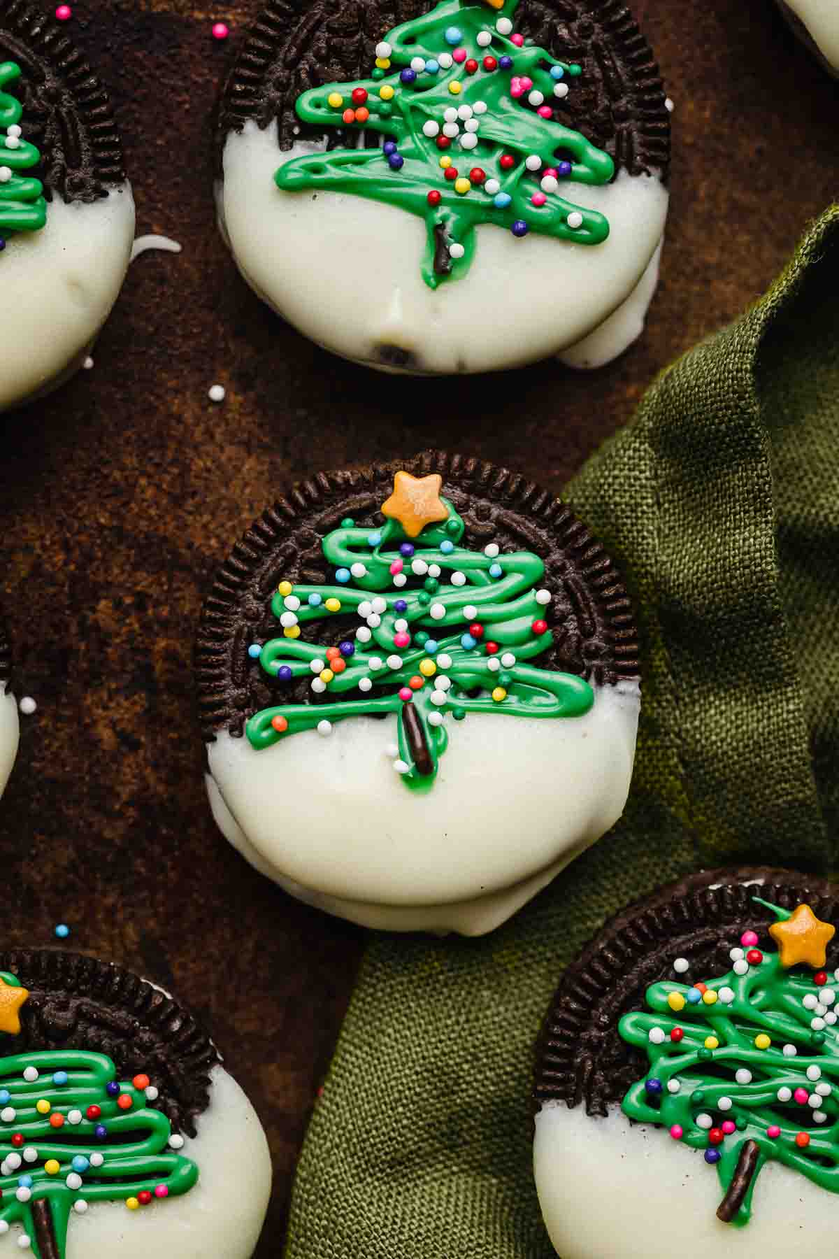Christmas Oreo on a green linen napkin, the Oreo is half dipped in white chocolate then has a green abstract christmas tree piped on the Oreo and sprinkled with nonpareil sprinkles and a star sprinkle on top.