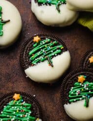Christmas Tree Oreos on a brown background, an Oreo half dipped in white chocolate with a green Christmas tree piped on the Oreos and topped with nonpareil sprinkles.