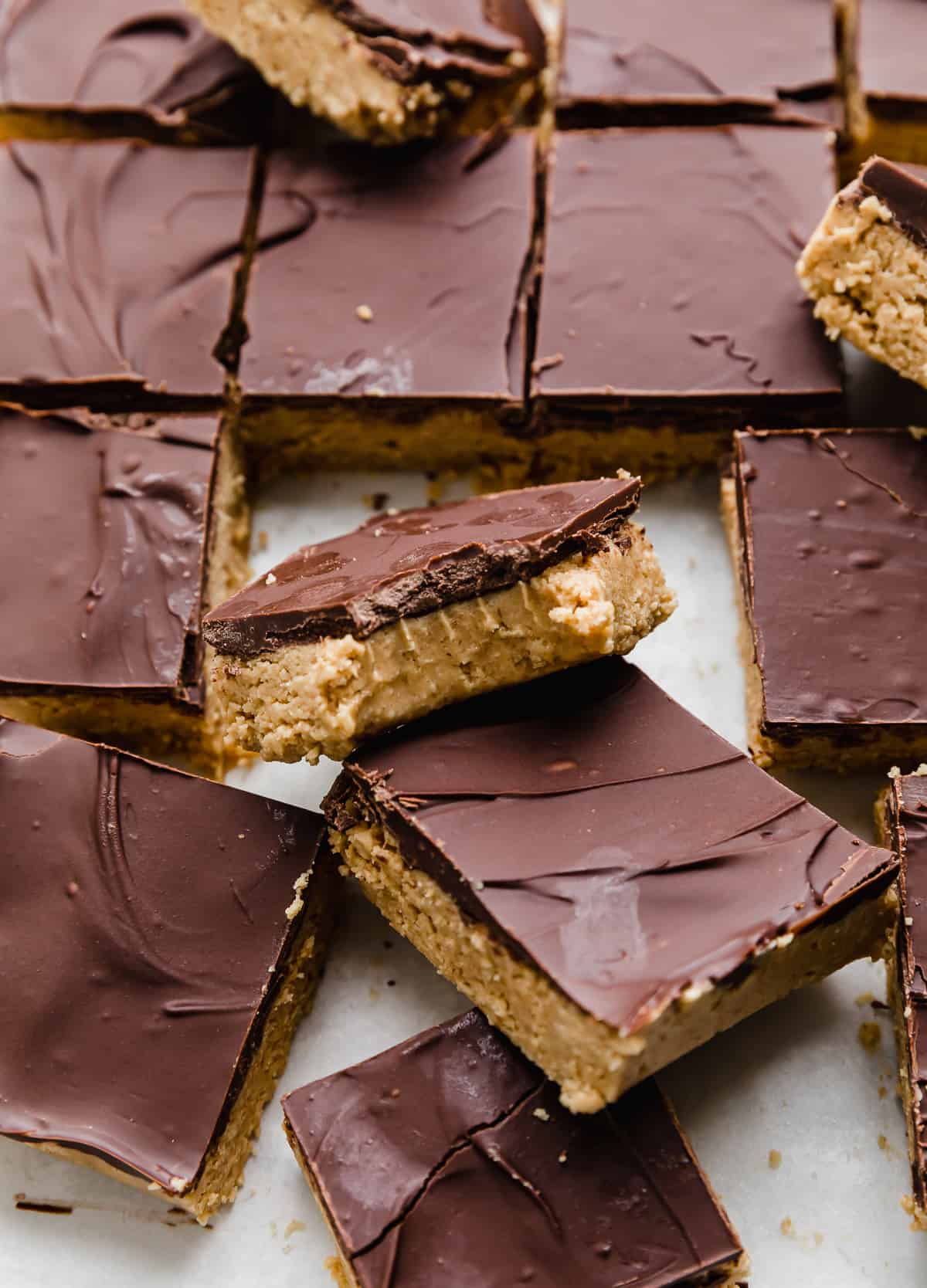 Chocolate Peanut Butter Bars cut into squares on a white background, with one no bake peanut butter bar topped with chocolate having a bite removed from it.
