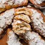 Flaky homemade Almond Croissants cut in half and surrounded by powdered sugar dusted Almond Croissants.