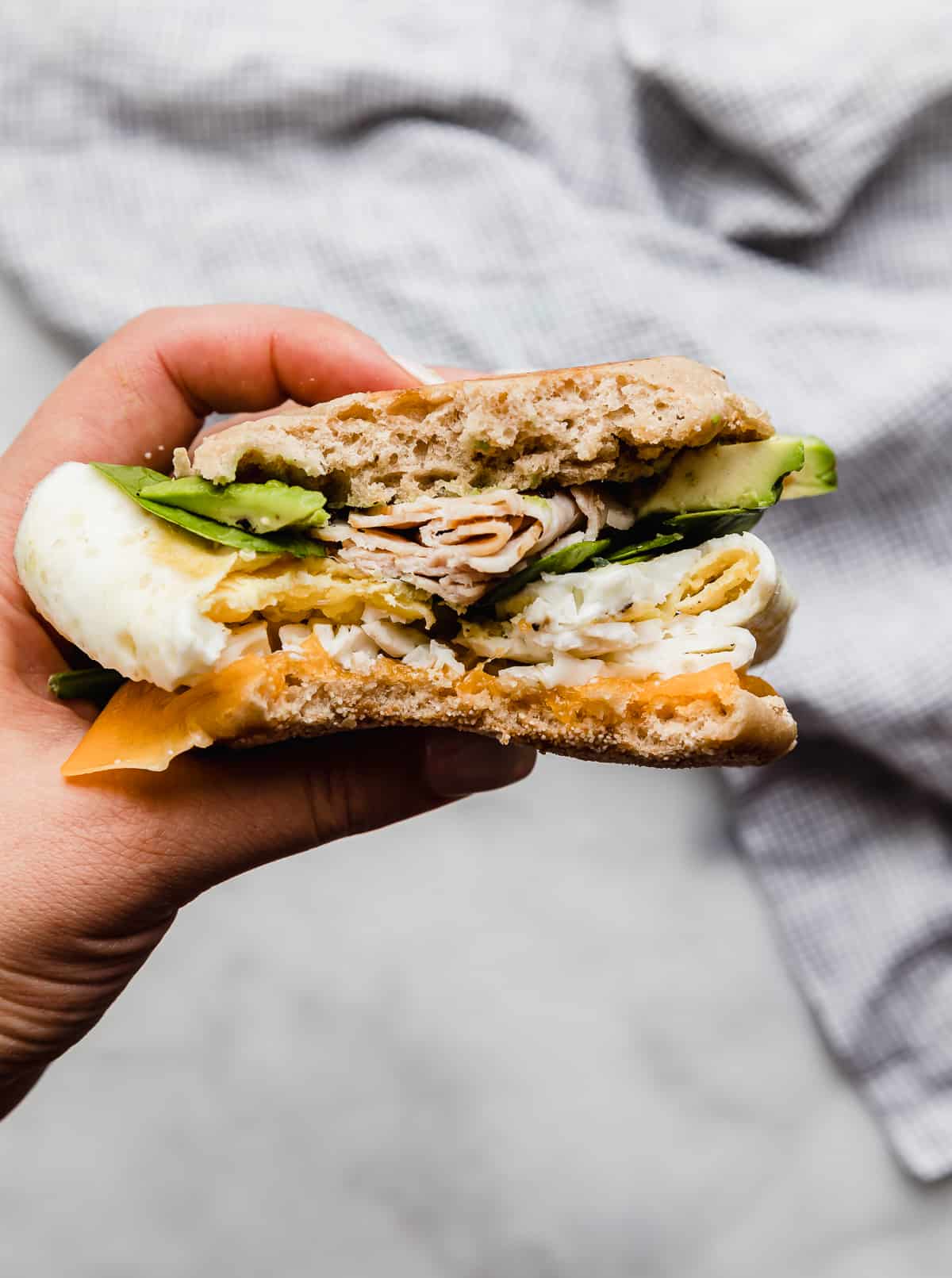 A hand holding a Turkey Breakfast Sandwich on an English muffin topped with avocado, spinach, turkey, eggs, and cheese.