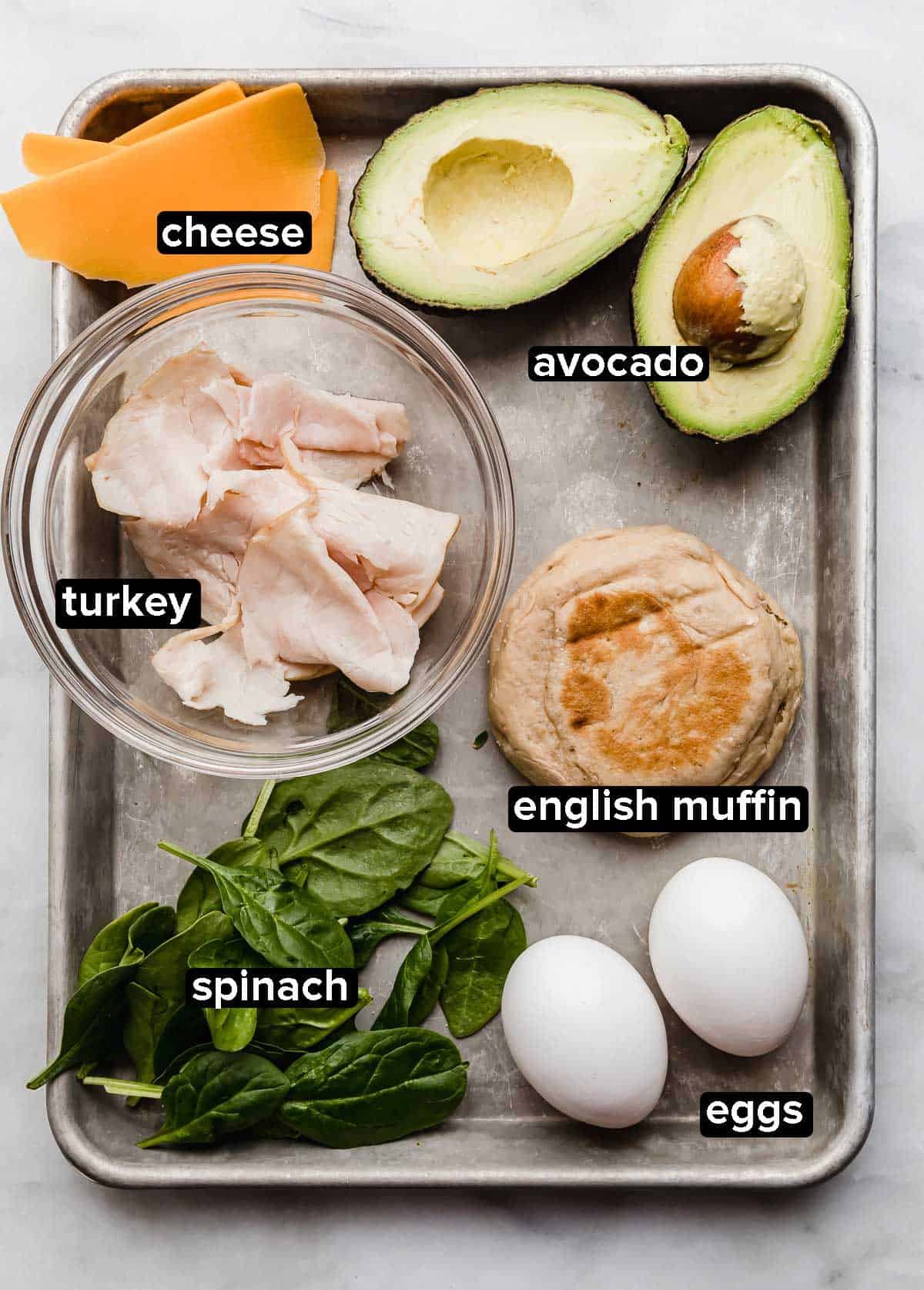 Turkey Breakfast Sandwich ingredients on a baking sheet: deli turkey meat, spinach, avocado, cheese, eggs, and English muffin.