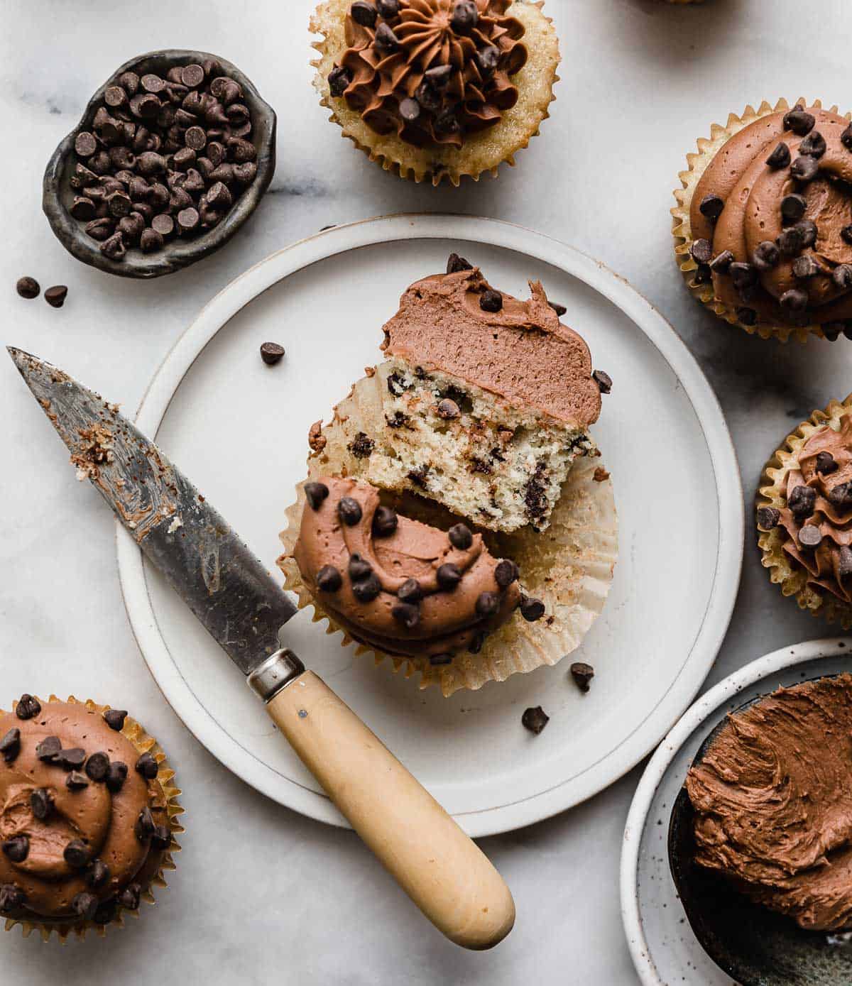 A Chocolate Chip Cupcake on a white plate, cut in half with a knife to the left of the plate.