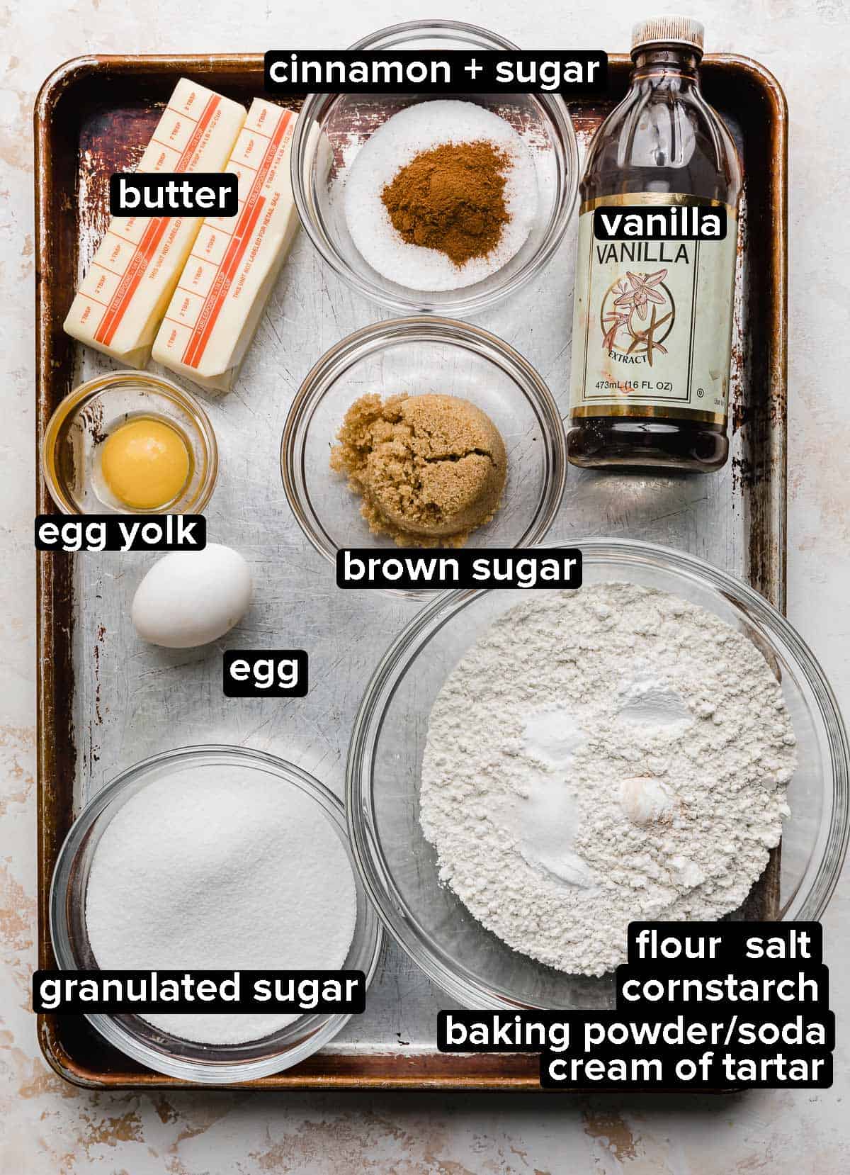 Snickerdoodle Bar ingredients on a baking sheet, each ingredients portioned into glass bowls: butter, egg, yolk, sugar, cinnamon, vanilla, flour, cream of tartar, and leavening agents.