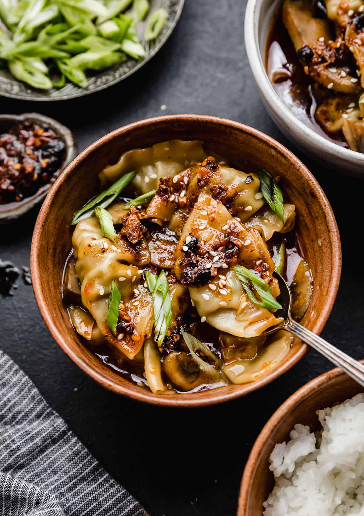 A brown bowl filled with Spicy Chicken Wontons topped with sliced green onions and chili sauce.