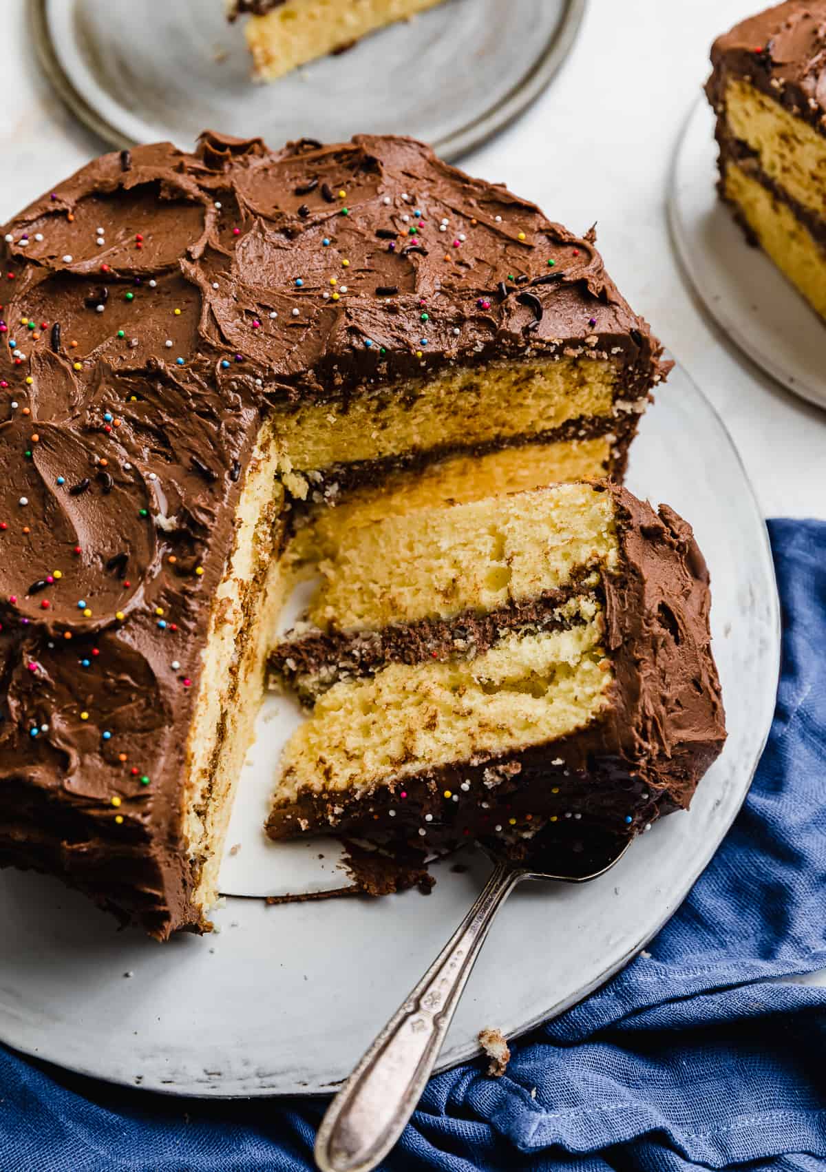 Yellow layer cake with chocolate frosting on a white cake plate, with a slice of cake cut and laying flat on the platter.