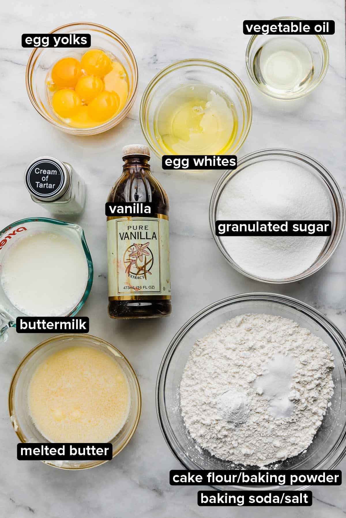 Ingredients used to make the best Yellow Cake with Chocolate Frosting recipe portioned into glass bowls on a white marble background.