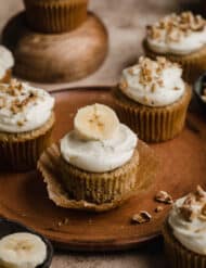 Banana Cupcakes topped with cream cheese frosting and a banana slice, on a brown plate.