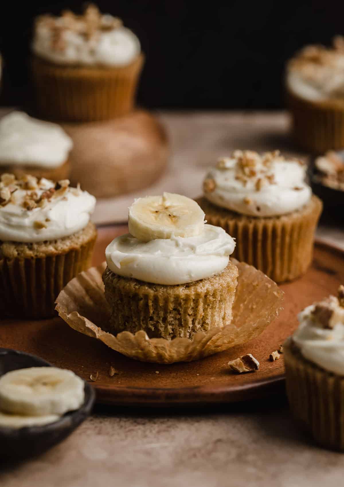 Banana Cupcakes on a brown plate against a dark brown background, with some of the cupcakes topped with chopped walnuts. 