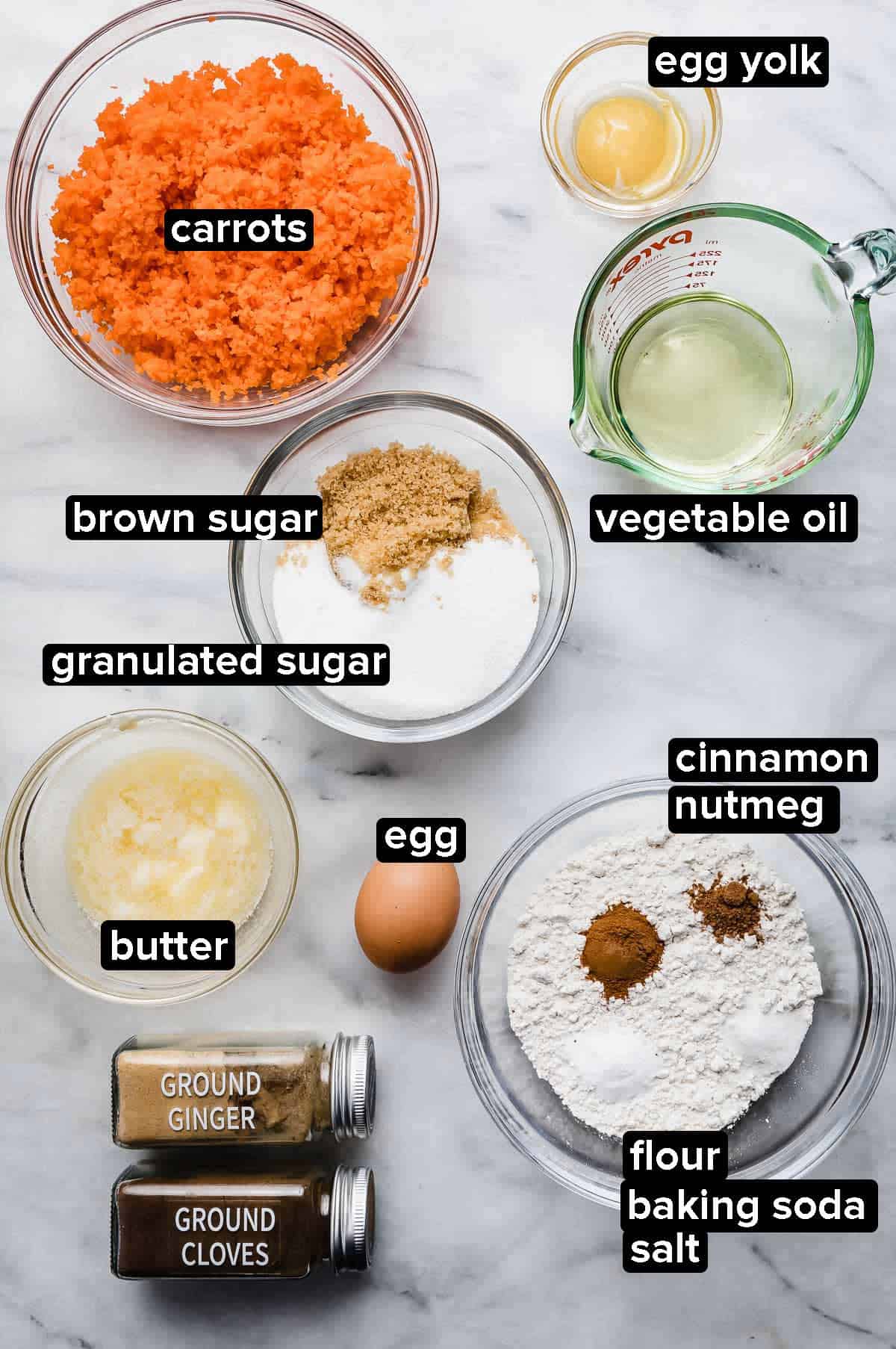 Carrot Coffee Cake ingredients portioned into glass bowls on a white marble background, shredded carrots, oil, sugars, melted butter, flour, eggs, cinnamon nutmeg, cloves, and ginger.