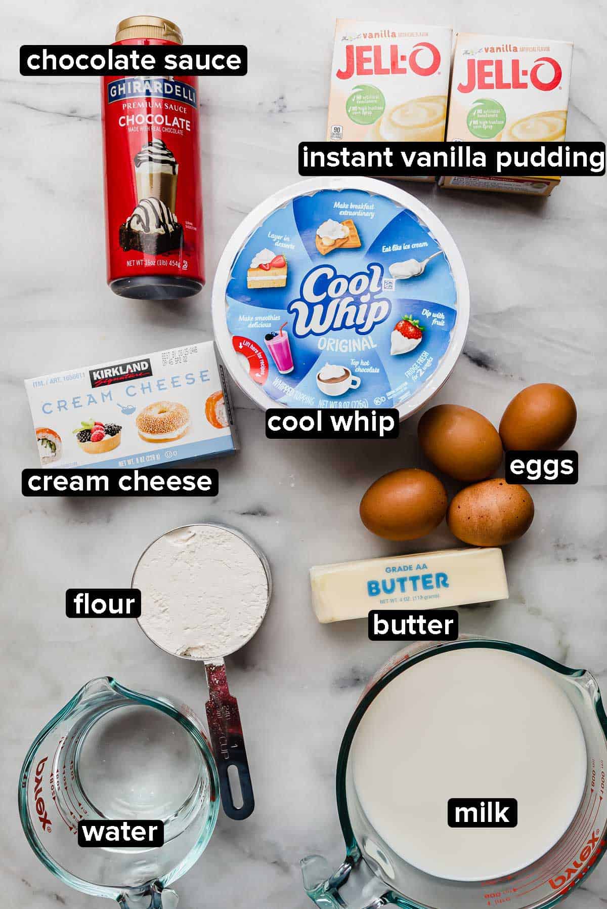 Cream Puff Cake recipe ingredients on a white marble table: instant vanilla pudding packets, cool whip, cream cheese, eggs, milk, water, butter, flour, and chocolate syrup.